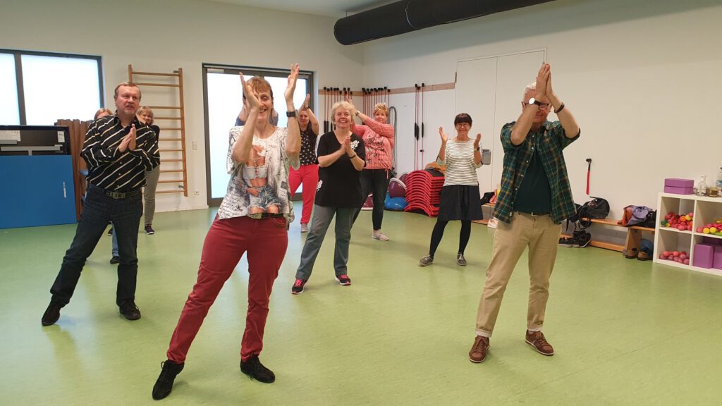 Die Linedance Gruppe beim Training im Sportraum. Der rechte Fuß ist ausgestreckt und sie klatschen die Hände über dem Kopf zusammen.