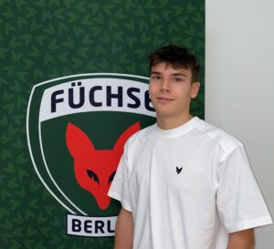 Auf dem Bild sieht man Konstantin Scholz vor dem Füchse Logo stehen. Konstantin absolviert ein duales Studium bei den Füchsen. Er ist vorallem im Bereich Fitter Fuchs zuständig.